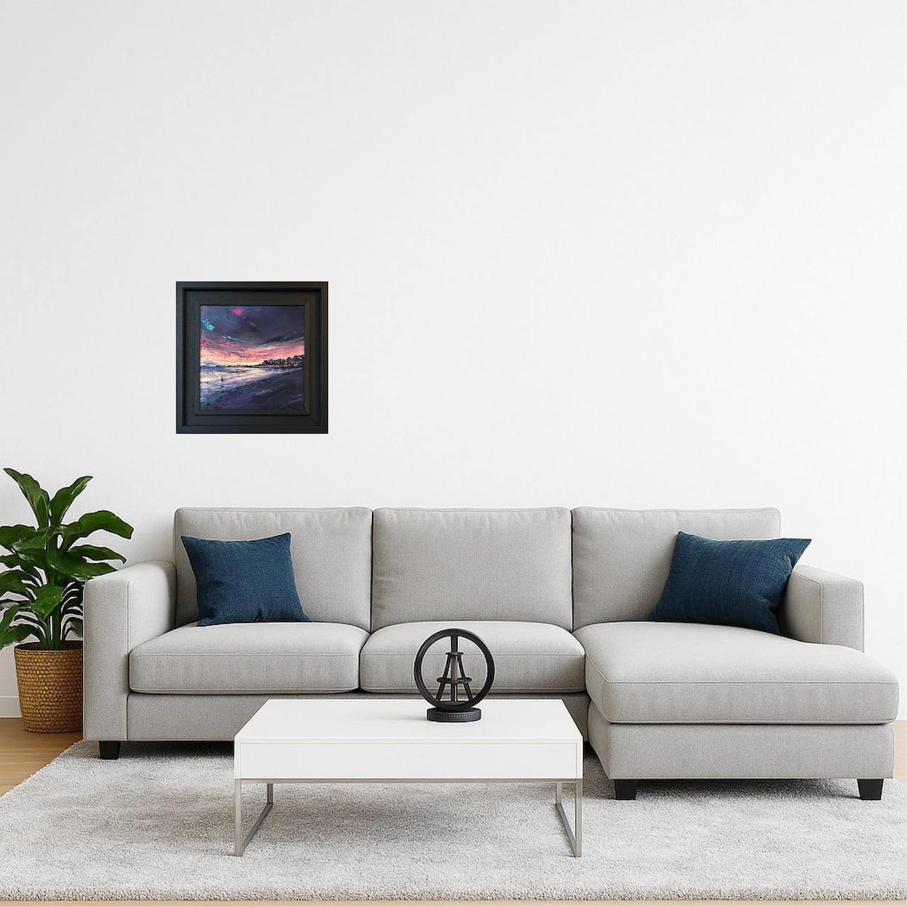 A framed landscape painting featuring a purple dawn over water, hung on a white wall above a modern light gray sectional sofa with two dark blue pillows in a styled living room setting.