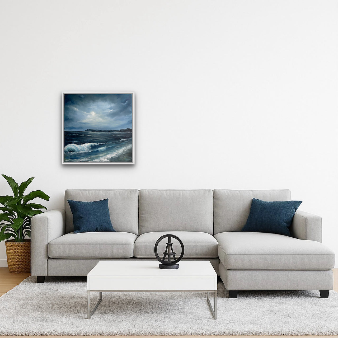 A framed square art print depicting a dramatic moonlit seascape with dark blue waves and a cloudy sky, displayed on a white wall above a modern light gray sectional sofa with navy blue pillows in a styled living room.