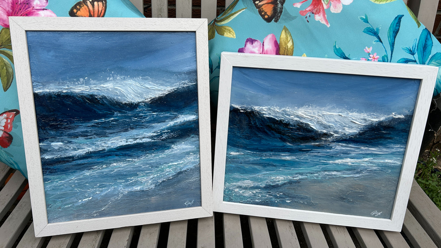 Two framed paintings of ocean waves on a wooden surface with a floral cushion in the background.
