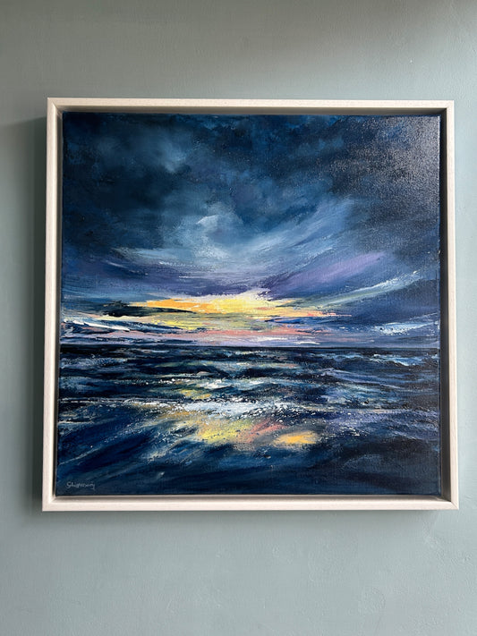 A framed square acrylic painting of a dramatic ocean sunset, featuring deep blue and navy waters leaning to purple with a bright yellow and pink horizon under a dark cloudy sky, mounted on a light gray wall.