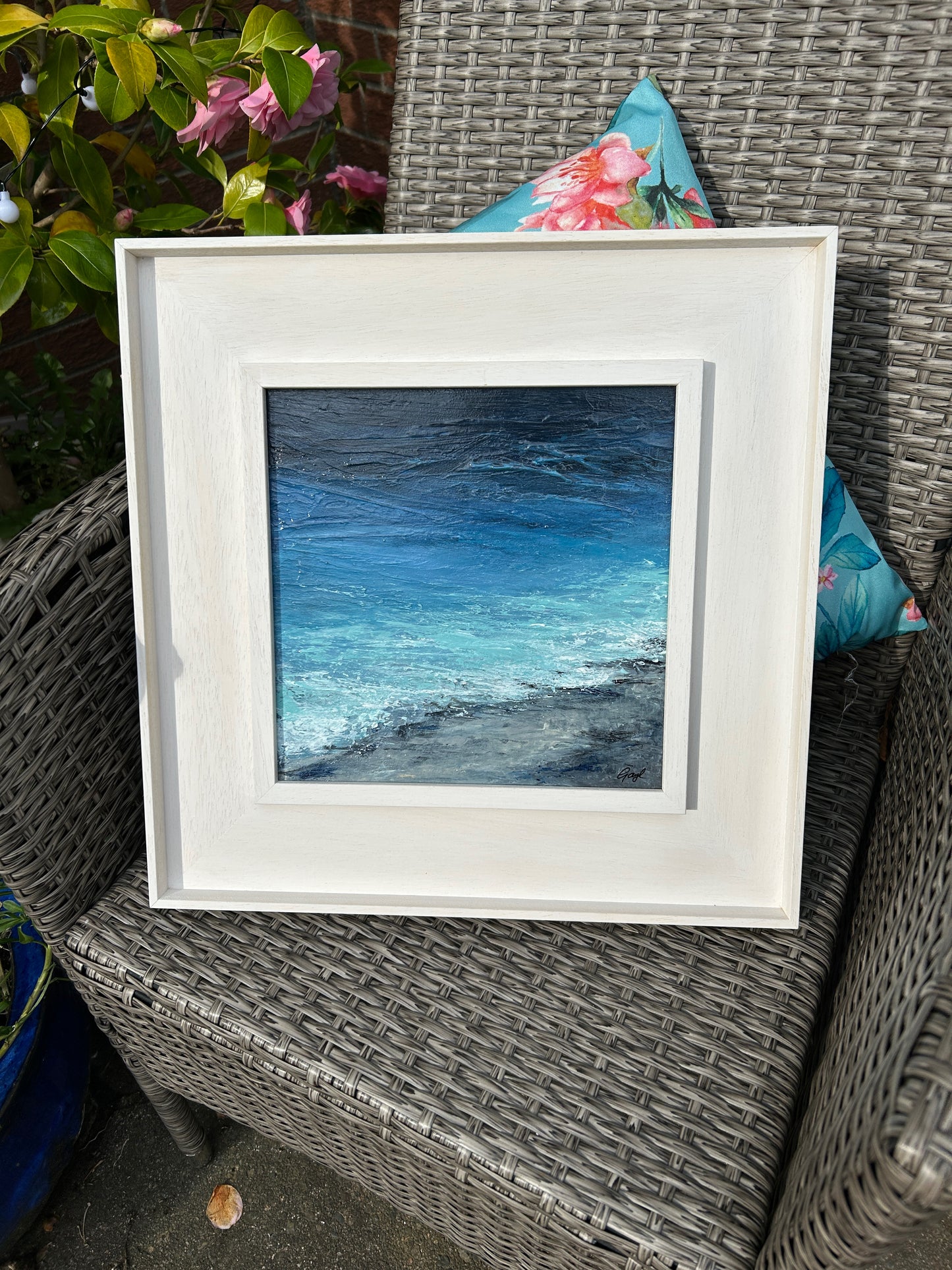 Framed artwork of a blue landscape on a wicker chair with floral cushions.