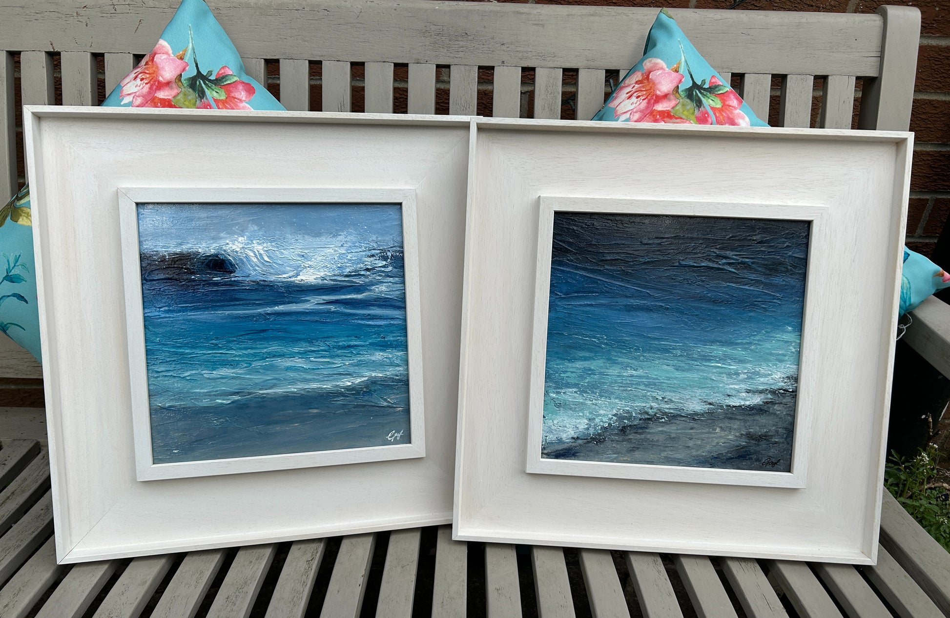 Two framed artworks of ocean scenes on a wooden bench with floral cushions.