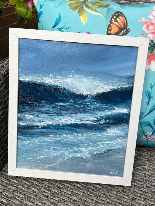 Framed artwork of a wave on a wicker chair with a floral pillow.
