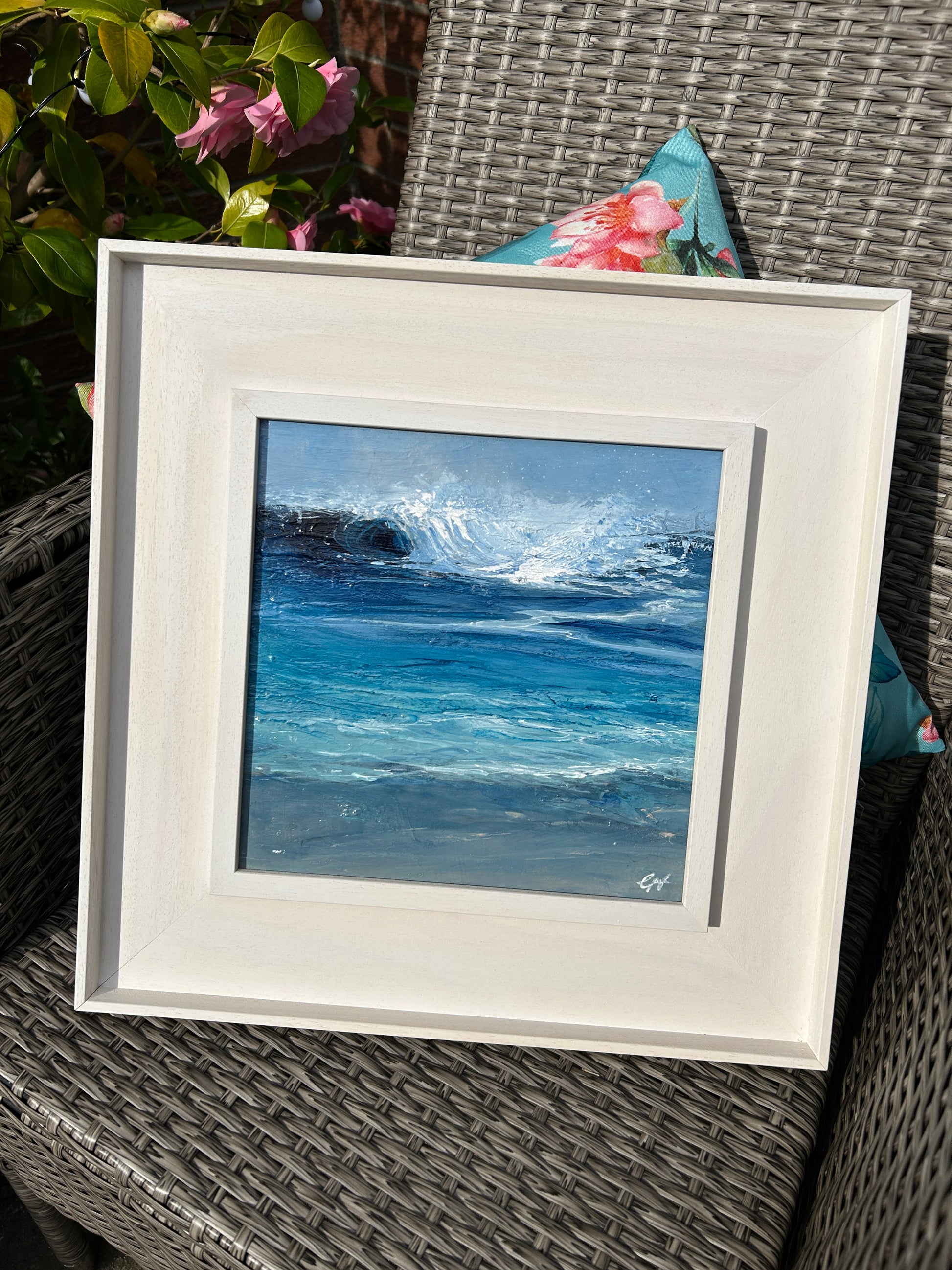 Framed artwork of a blue ocean scene on a wicker chair with floral cushions.