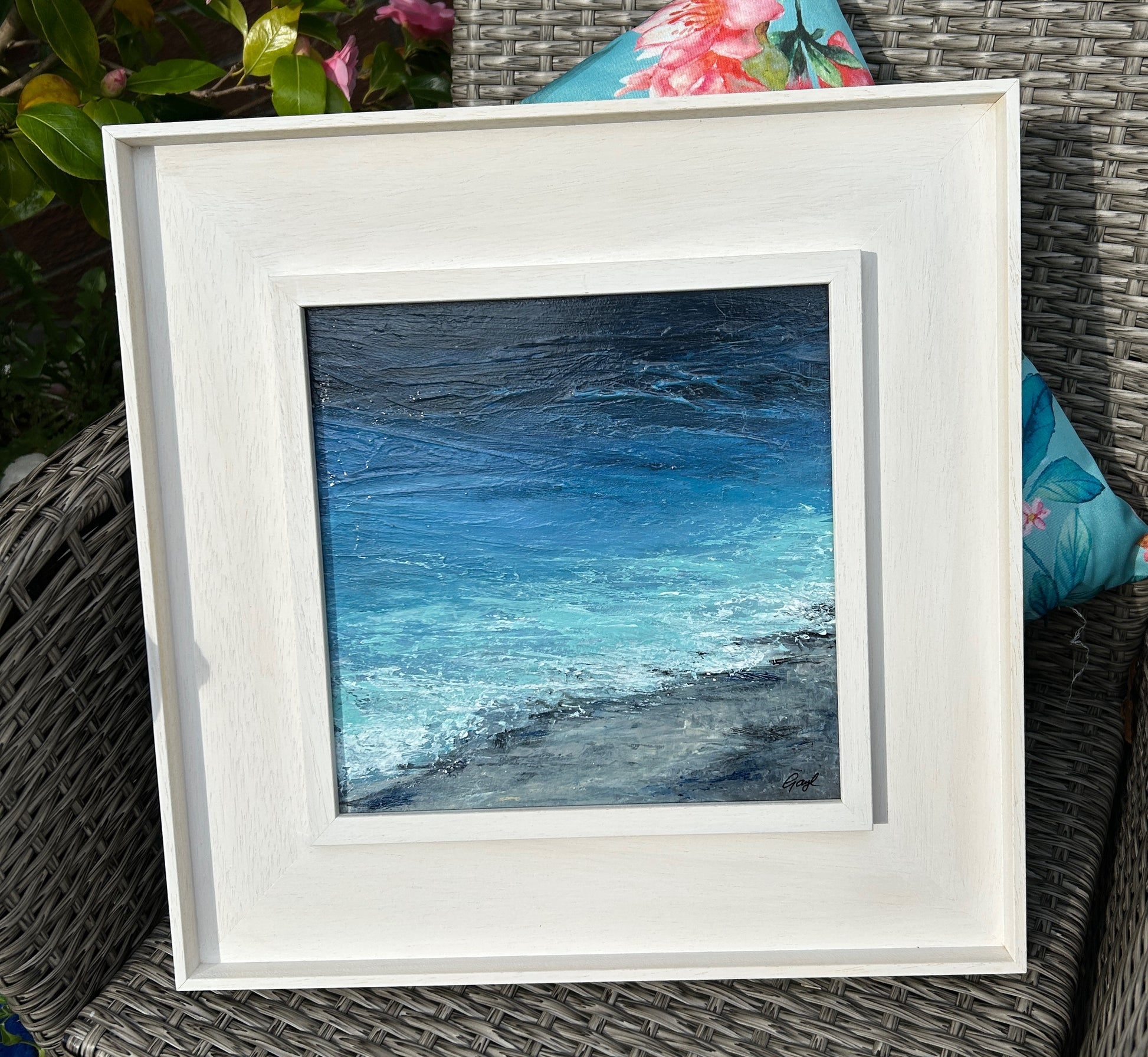 Framed artwork of a blue ocean scene on a wicker chair with floral cushions.