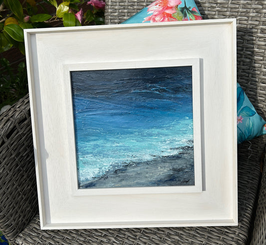 Framed artwork of a blue ocean scene on a wicker chair with floral cushions.