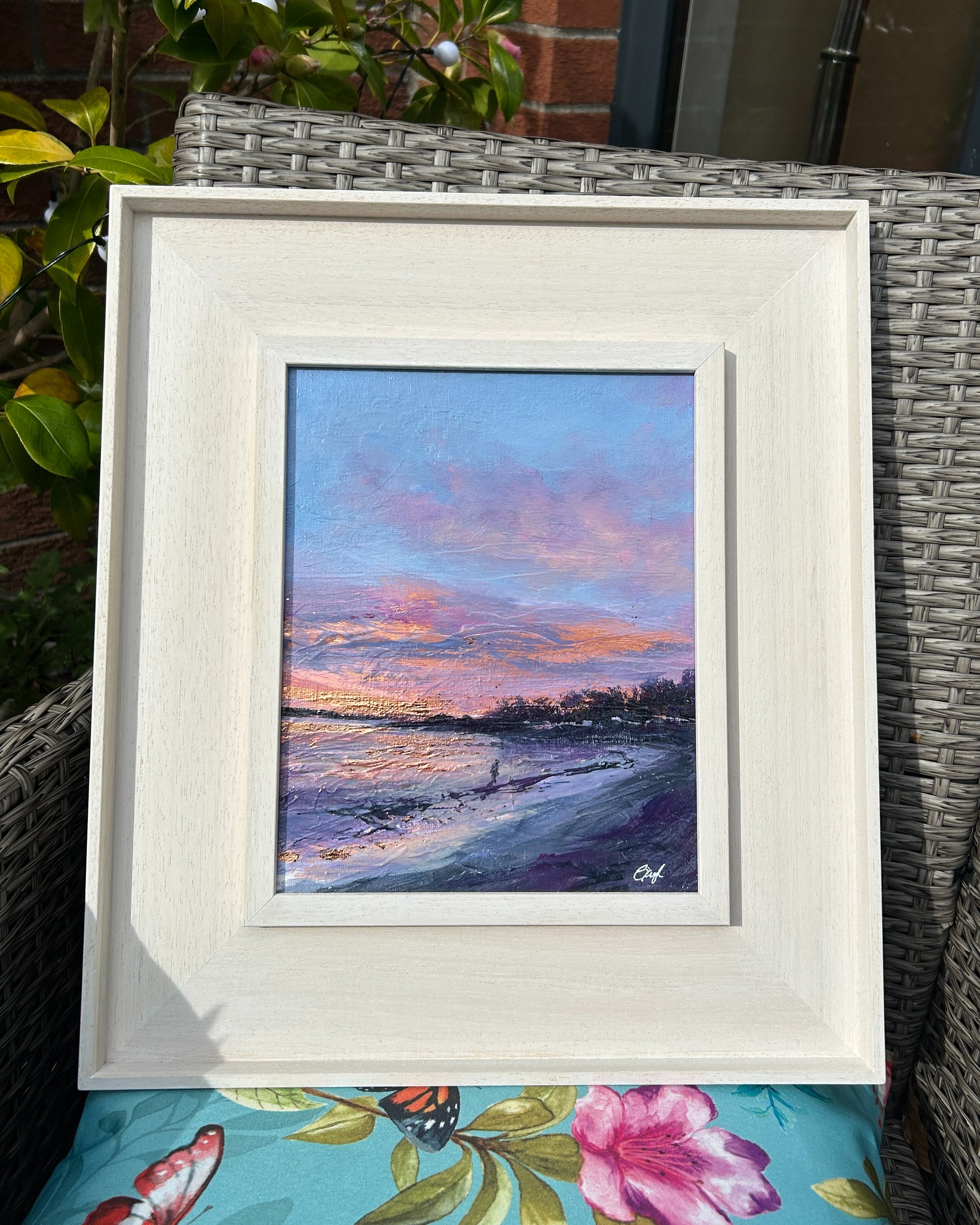 Framed artwork of a beach scene at dawn, placed on a textured surface with floral patterns.