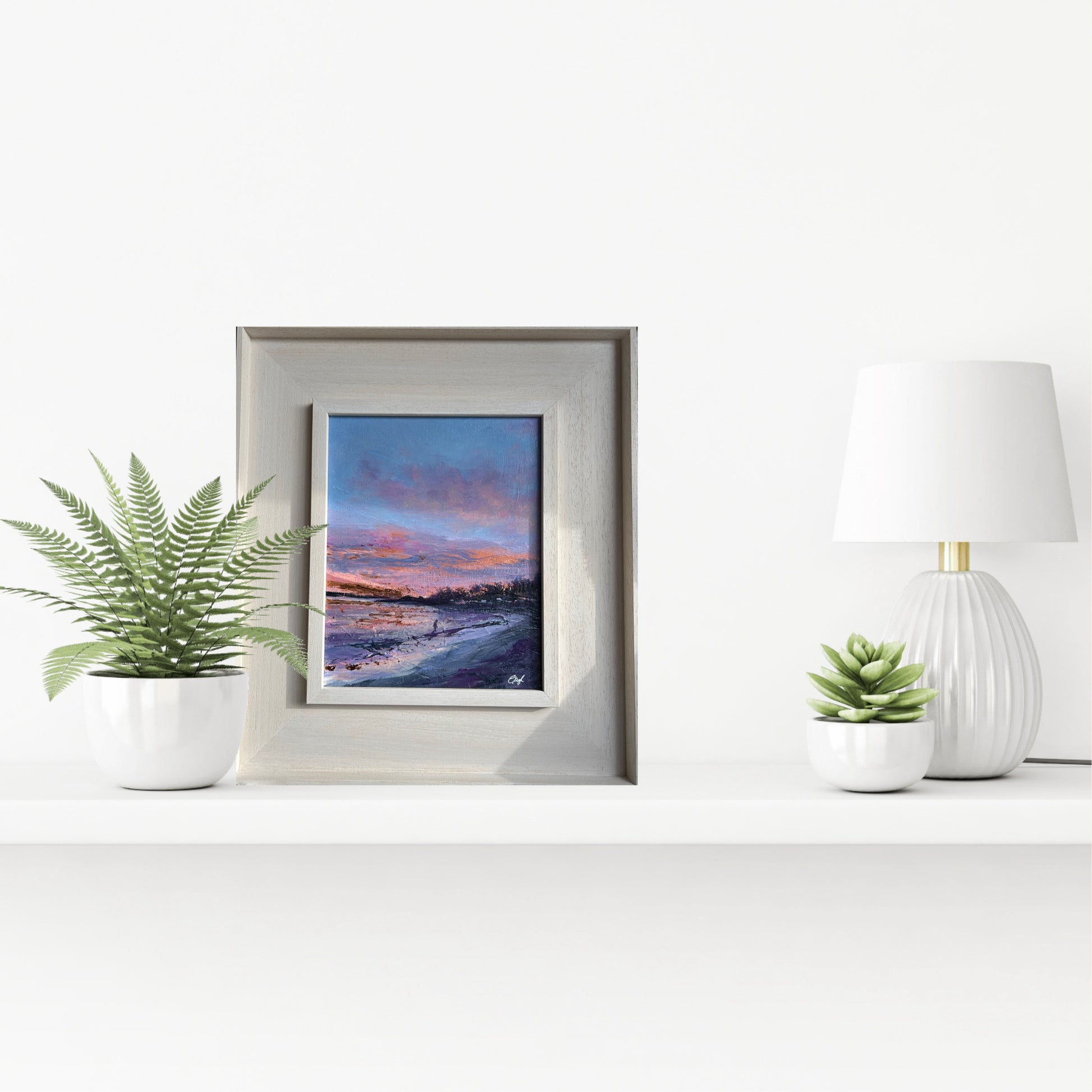 Framed artwork on a white surface with plants and a lamp. Helens Bay dawn sea swimming