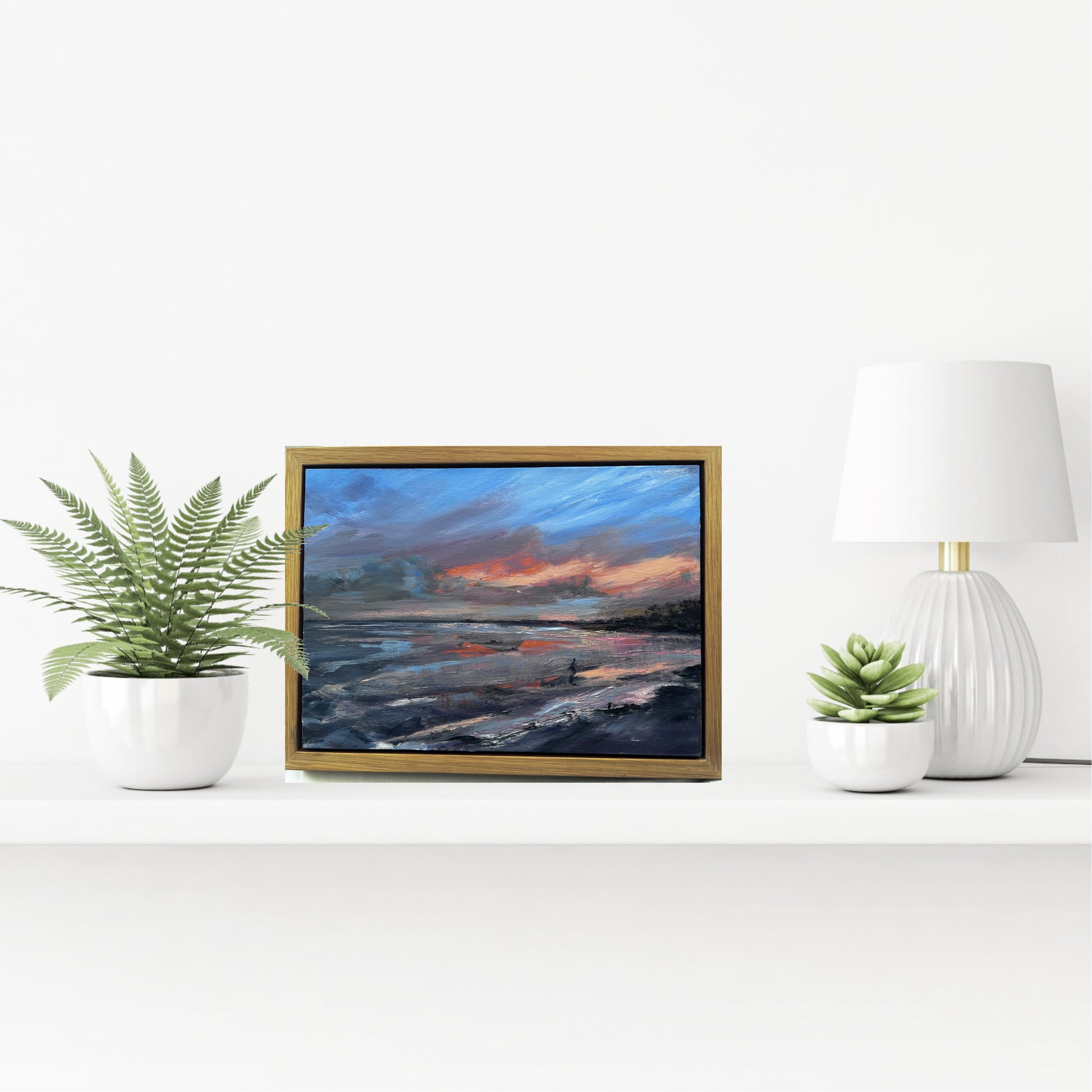 Framed artwork of a seascape with a sunrise over water, placed on a white surface with decorative plants and a lamp. Sea swimming at Helens Bay