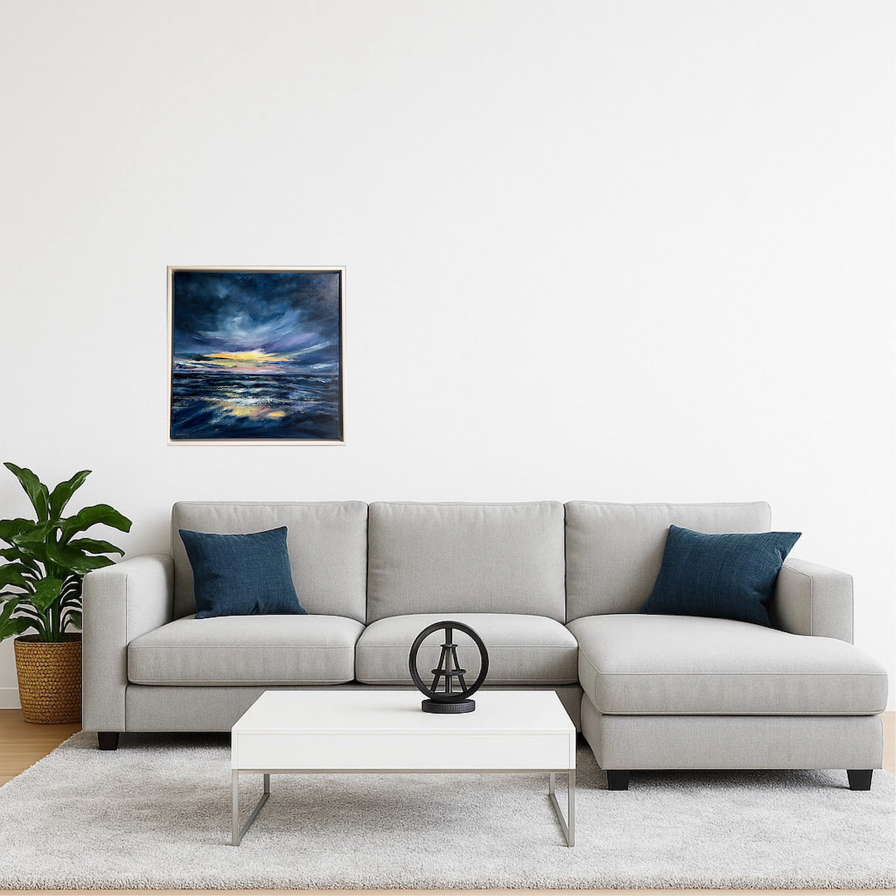 A modern living room setup featuring a light gray sectional sofa with two dark blue throw pillows, a white coffee table, and a framed square canvas print depicting a dramatic stormy seascape hanging on the wall.