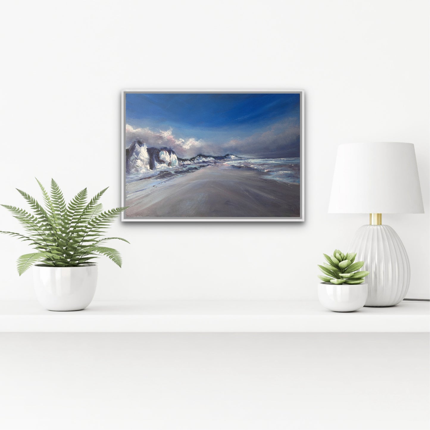 Framed beach scene on a wall with decorative plants and a lamp.